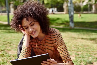 Gün boyunca parkta tek başına otururken tabletten kitap okuyan gülümseyen siyah bir kadın.