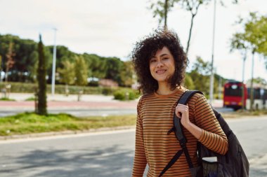 Sırt çantasıyla gün boyunca tek başına yürürken kameraya bakan gülümseyen genç bir kadın.