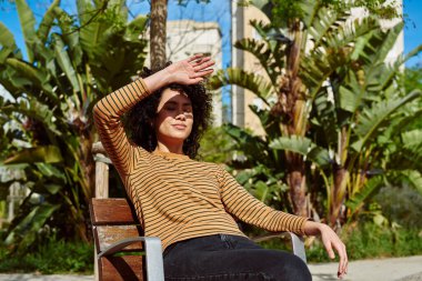 Güzel, genç, siyah bir kadın gün boyunca dışarıda bir sandalyede dinlenirken yüzünü güneşten koruyor.