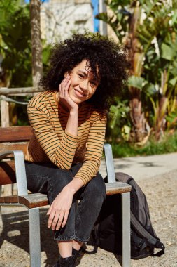 Güneşli bir günde, açık havada bir sandalyede tek başına oturmuş kameraya bakan gülümseyen siyah bir kadın.