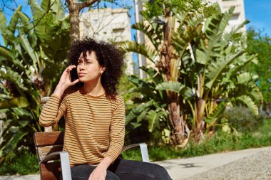 Güneşli bir günde, açık havada bir sandalyede tek başına oturan sevimli siyahi kadın bir telefon görüşmesi yapıyor.