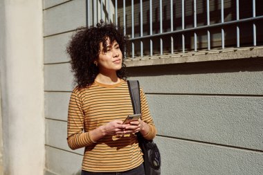 Şirin, genç, siyah bir kadın, elinde cep telefonu, elinde sırt çantasıyla, gün boyu dışarı bakarken, dikkatle başka tarafa bakıyor.