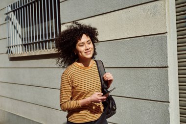 Sırt çantası ve cep telefonuyla gün boyunca gülümseyen siyah bir kadın.