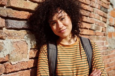 Sırt çantasıyla tuğladan duvara yaslanmış kameraya bakan sevimli siyah bir kadın.