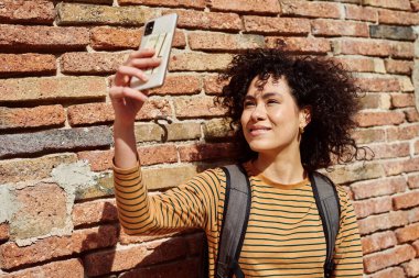 Gülümseyen genç siyahi kadın, gün boyunca dışarıda tuğla bir duvara yaslanırken selfie çekiyor.