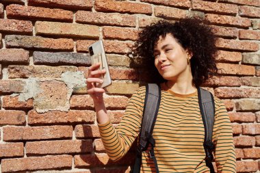 Gün boyunca dışarıda tuğladan bir duvarın önünde durarak selfie çeken sevimli genç siyahi kadın.