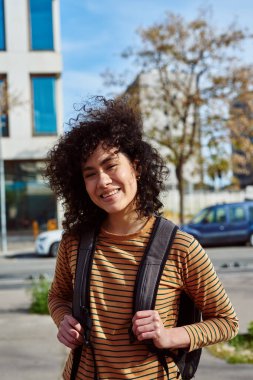 Sırt çantalı mutlu siyah bir kadın. Gün boyunca şehirde tek başına dikilirken kameraya gülümsüyor.