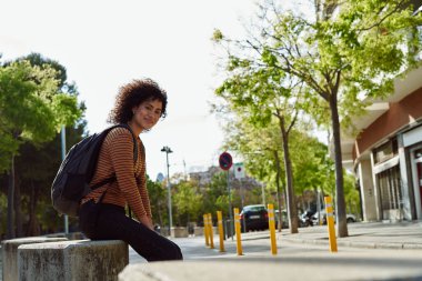 Sırt çantalı neşeli siyah bir kadın kameraya gülümserken gün boyunca dışarıda taş bir levhada tek başına oturuyor.