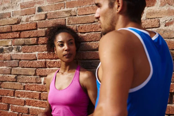 Athletic young couple having a chat while standing against a brick wall outdoors during the day