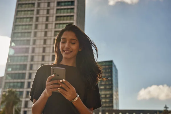 Young laughing businesswoman standing and text messaging in front of building