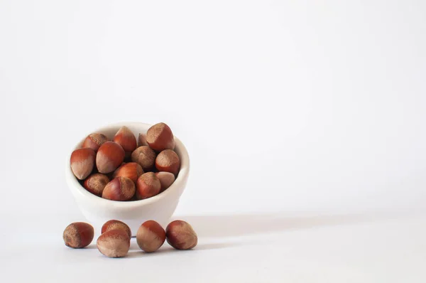 Hazelnuts, heap or stack of full hazelnuts in white bowl and some on white background. Healthy and tasty vegan food, isolated and selective focus.