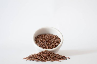 Keten tohumları, küçük beyaz bir kaseden dökülen tohumların profil görüntüsü izole edilmiş arka plandaki minimalist bir sahnede beyaz bir yüzeye dökülüyor..