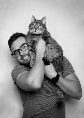 Kollarında tekir kedi tutan sakallı ve gözlüklü gülümseyen genç bir adamın stüdyo portresi. Gri arka plan. Beyaz arka plan. Hayvan sevgisi konsepti.