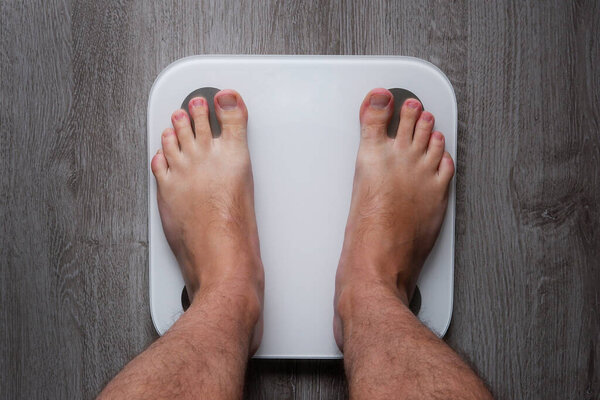 Top view on the feet of a barefoot man standing on a smart scale. White smart weights with empty balance standing on a gray wooden floor. Conceptual photo of weight loss.