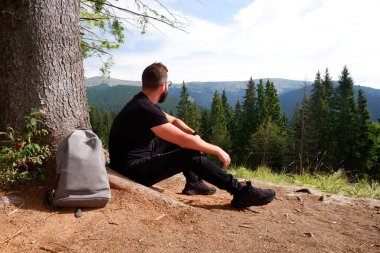 A young man in black clothes and a backpack sits alone outdoors with mountains and a forest in the background. Travel concept.