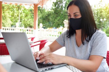 Tıbbi maskeli bir kız dizüstü bilgisayarla video konferansı yapıyor ve dışarıda bir kafede oturuyor. Çevrimiçi eğitim ve çalışma mesafesi. Koronavirüs Pandemik Karantina