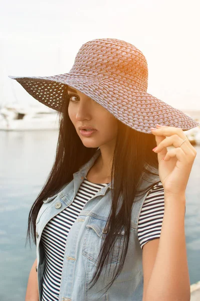 Kot yelekli, esmer, güzel bir kadının portresi Kafkasya ve bir elinde hasır bir şapka tutan denizin arka planında çizgili bir elbise. Yatlı rıhtım.