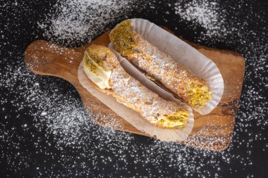 Top view of two fresh cannoli with pistachios in paper on a cupboard on a black background powdered with sugar