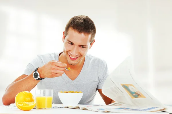 Glücklicher junger Mann, der Zeitung liest, während er energisch frühstückt — Stockfoto