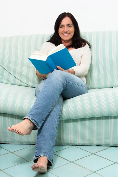 Donna dai capelli scuri sdraiata sul divano e che legge un libro — Foto Stock