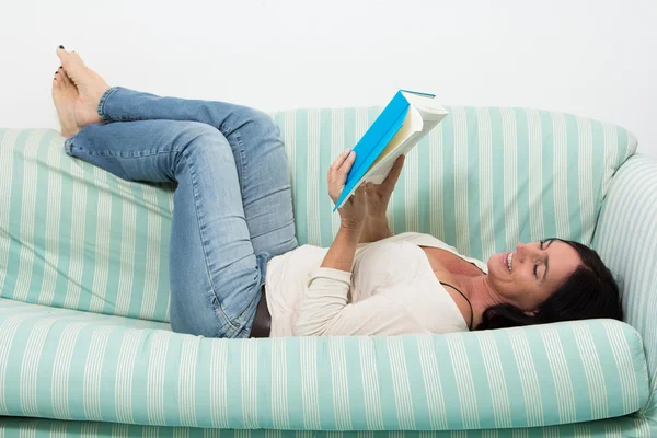 Donna dai capelli scuri sdraiata sul divano e che legge un libro — Foto Stock