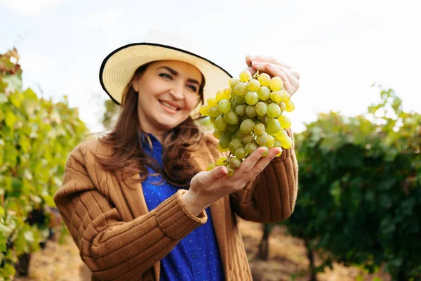 Winzer lächelt erwachsene Frau mit Hut, die einen Bund weißer Trauben in der Hand hält — Stockfoto