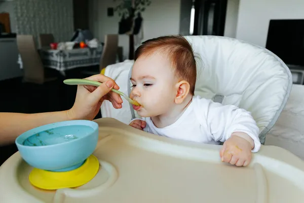 Mãe americana alimentando um bebê com uma colher, no fundo da sala de estar. — Fotografia de Stock