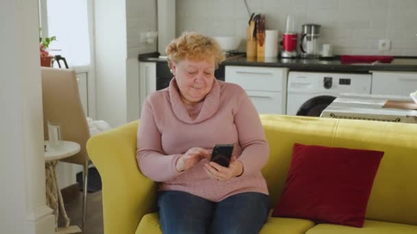 Senior lady za pomocą smartfona surfing w sieci siedzieć na kanapie w domu w salonie, — Wideo stockowe