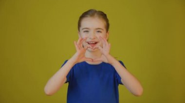 Çocuk kameraya bakıyor ve elleriyle kalp işareti gösteriyor.,