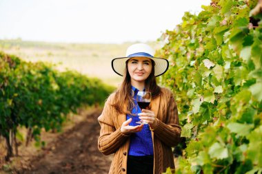 Çekici şarap üreticisi kadın elinde bir bardak kırmızı şarapla kameraya bakıyor.