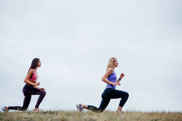 Siyah spor giyimli iki genç atletik kadının yan görüntüsü, dışarıda esneme egzersizleri yapıyor. — Stok fotoğraf