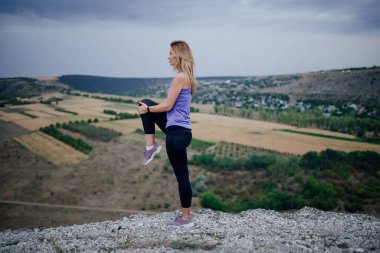 Siyah spor kıyafetli genç bir kadın gökyüzü arka planında esneme egzersizleri yapıyor..