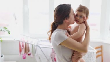 Tatmin olmuş genç bir anne evdeki büyük pencerede bebeğini öpüyor ve sarılıyor..