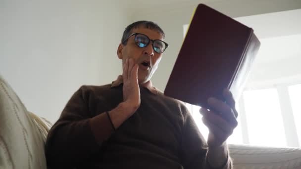 Старый индеец читает книгу дома в очках, испуганных от шока с неожиданным лицом — стоковое видео