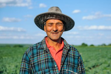 Kafasında şapka olan ekose gömlekli bir erkek çiftçinin portresi tarım tarlasından memnun. Güzel bir çalışmadan sonra kameraya bakan yaşlı çiftçinin yavaş hareket eden portresi..