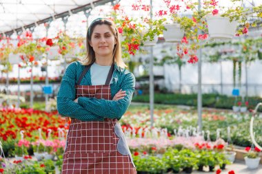 Çekici, mutlu, önlüklü, kollarını kavuşturup gülümseyen genç bir kadın. Etrafında bir sürü çiçekle bir serada dikilirken. Boşluğu kopyala