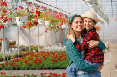 Kırmızı kare gömlekli ve şapkalı neşeli bir kız çocuğunun annesinin ellerinde oturması. Mutlu güzel kız ve anne ailesi kameraya bakıp serada poz veriyor. Bahçıvan ailesi, kopyalama alanı