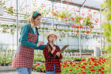Güzel kadın anne kızıyla birlikte çiçeklerle ve ev bitkileriyle dolu serada çalışıyor. Elinde not defteri olan kız bitkiler, kopyalama alanı hakkında bilgi yazar