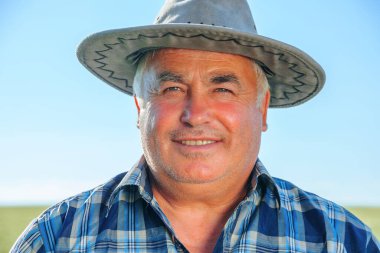 Güneşli bir yaz günü kırsal kesimde tarım çiftliğinde dikilirken kameraya bakan şapkalı yaşlı, mutlu erkek işçi kırsal kesimde mutlu, olgun çiftçi.