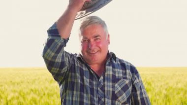 Dost canlısı, kareli gömlekli bir adam kameraya bakıyor ve kırsal kesimde güneş ışığında tarım alanında çalışırken şapka takıyor.