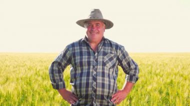 Kareli gömlekli ve şapkalı mutlu bir adam elini beline koyuyor ve tarımsal alanda dikilirken gülümseyerek kameraya bakıyor..