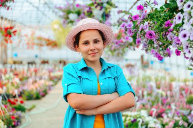Front view of a young agronomist girl in a greenhouse, the woman crosses her arms over her chest and smiles playfully. The agronomist girl stands in a hat and shirt among the flowers.
