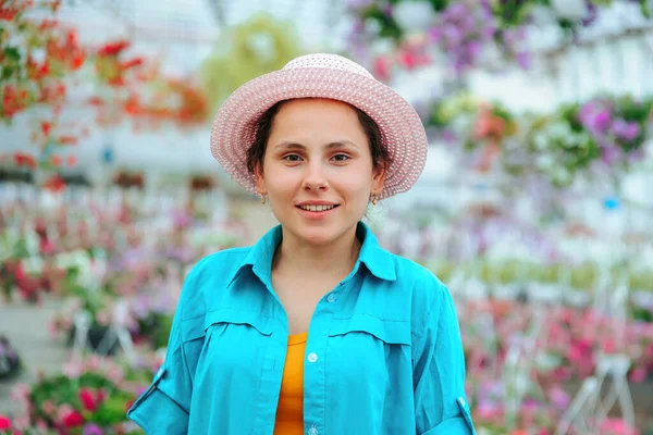 Young woman nice florist stands in a greenhouse and looks into the camera. The woman turned her head to the right. Girl smiles calmly. The agronomist is dressed in a blue shirt and has a hat on had.