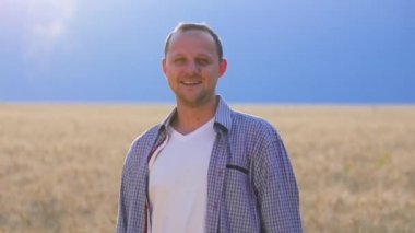 Front view of a young male farmer against the background of a wheat field and looking at camera, clear sky stands and smiles. The guy is happy with his success. The agronomist is slightly unshaven.