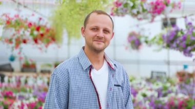 A young attractive farmer stands in a greenhouse and looking at camera. The head is slightly tilted to the left. Man smiles a calm smile. The agronomist is dressed in a plaid shirt. Copy space