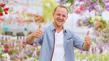 A very happy lucky man stands in a greenhouse and shows thump up with both hands. The businessman laughs heartily and shows how successful everything is. Dressed in a plaid shirt.