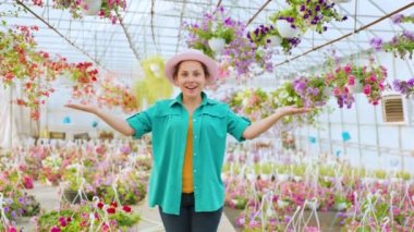 Standing in a greenhouse with indoor plants, young woman florist smiles happy and spreads her arms, showing the beauty of flowers. The girl is dressed in casual style, jeans, shirt, hat.