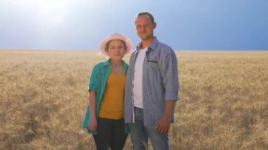 Front view of a pair of young farmers agronomists standing in the background of a wheat field during the day. A man and a woman hold hands, look around the field and look at each other.