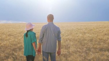 Rear view of a pair of young farmers agronomists standing in the background of a wheat field during the day. A man and a woman hold hands, look around the field and look at each other.
