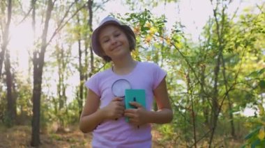 the portrait of a small child naturalist botanist with magnifying glass and notebook in hand, is in the forest. kid looking at the camera and smiling